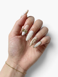 Main Cherry Rouge portant des faux ongles en amande nude avec détails dorés, nœud, perles et texture serpent