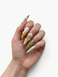 Main portant des faux ongles Cherry Rouge stiletto nude et parme avec fleurs 3D, détails dorés et ornements bijoux