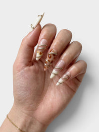 Main Cherry Rouge portant des faux ongles en amande Gilded Twist avec motifs torsadés dorés sur fond nude et crème