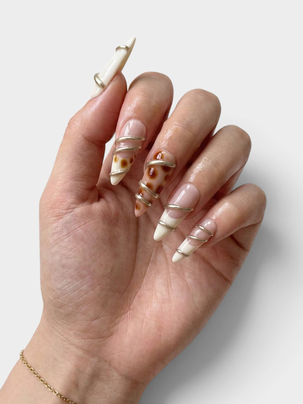 Main Cherry Rouge portant des faux ongles en amande Gilded Twist avec motifs torsadés dorés sur fond nude et crème