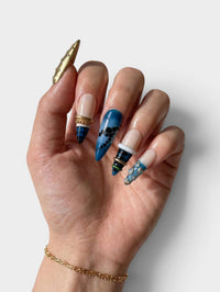 Main Cherry Rouge Gilded Cascade avec faux ongles en ABS bleu et nude ornés de bijoux dorés et de motifs géométriques