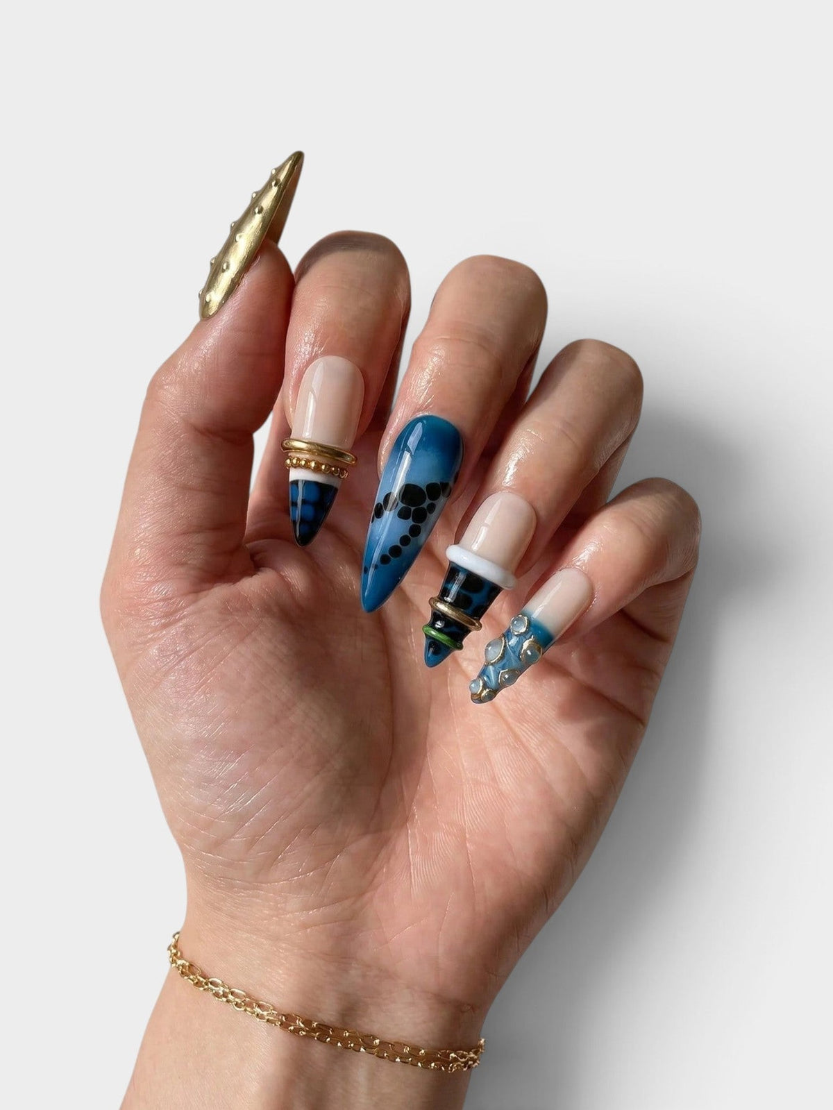Main Cherry Rouge Gilded Cascade avec faux ongles en ABS bleu et nude ornés de bijoux dorés et de motifs géométriques