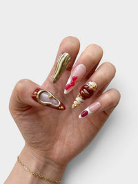 Manucure Cherry Rouge artistique en forme amande avec détails 3D dorés, effets texturés et motifs lèvres sur fond blanc